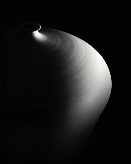 Monochromatic still life of a ceramic vase illuminated by a single light source on a stark black background, evoking a sense of artistic minimalism.