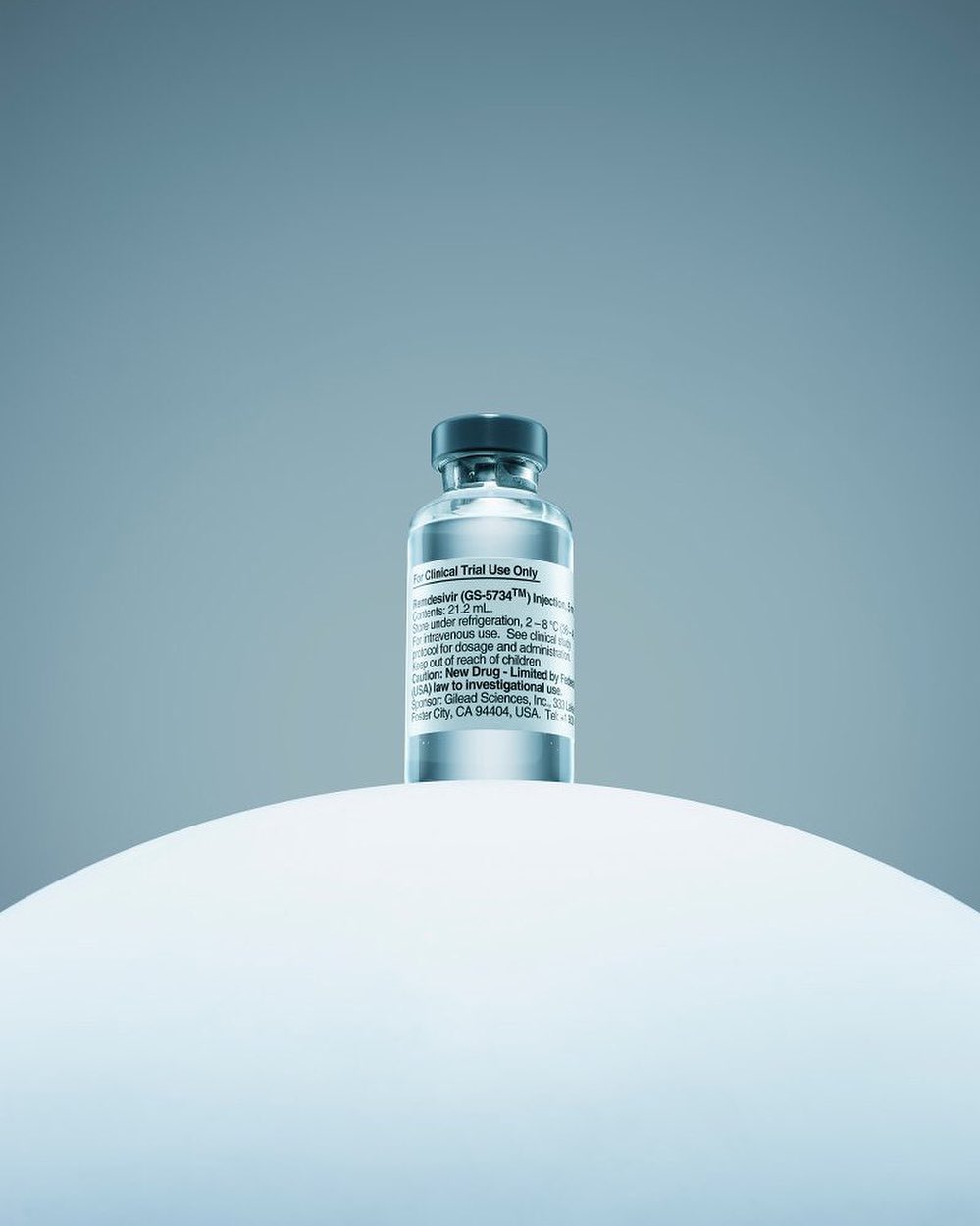 A close-up showcases a vial of Remdesivir atop a white, rounded surface against a soft, gradient blue backdrop, emphasizing its pharmaceutical nature.
