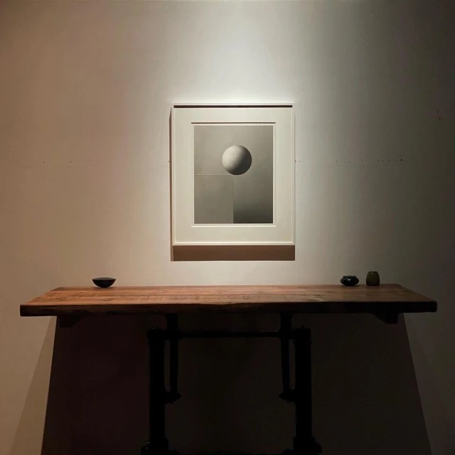 Artwork featuring a gray-scale sphere hangs above a wood table with small vessels against a neutral wall in a softly lit indoor setting.