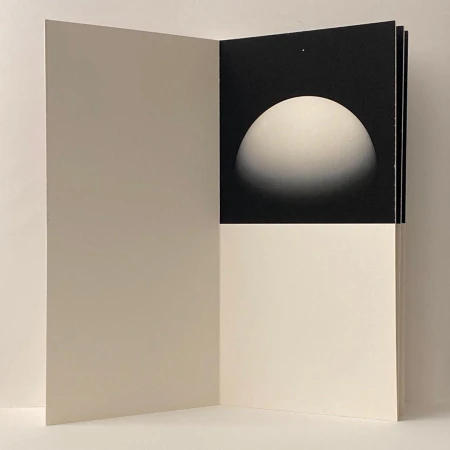 Open art book with a black page showing a white hemisphere graphic on the right and blank cream page on the left.