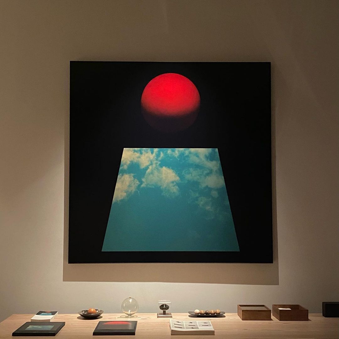 Abstract artwork features a red sphere above a trapezoid filled with a cloudy sky, presented on a beige wooden table in an indoor space.