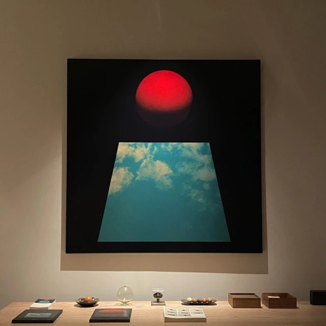 Abstract artwork features a red sphere above a trapezoid filled with a cloudy sky, presented on a beige wooden table in an indoor space.