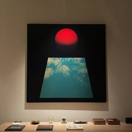 Abstract artwork features a red sphere above a trapezoid filled with a cloudy sky, presented on a beige wooden table in an indoor space.