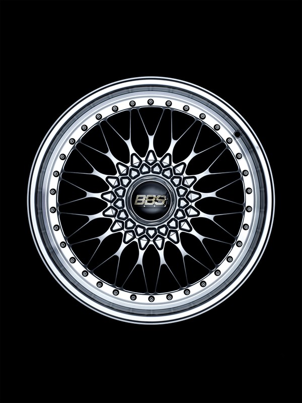 Detailed shot of a silver multi-spoke BBS wheel rim with a black center cap, captured in a dark, high-contrast studio setting.