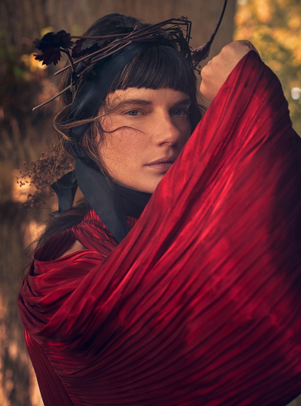 Portrait of a woman with a floral and twig crown, wearing a draped red dress, in a natural outdoor setting with an eye-level angle.