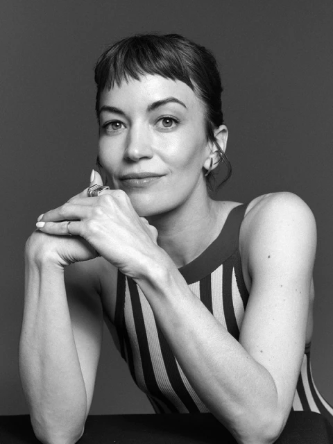 Monochrome portrait of a woman with short brown hair and striped top leaning her head on her hands, set against a grey backdrop.