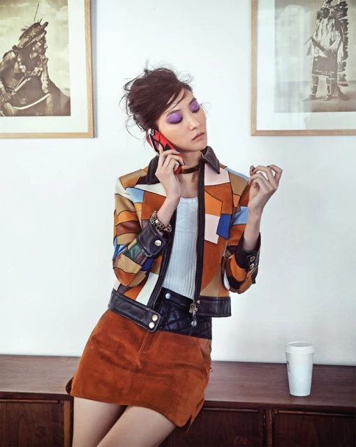 A young adult woman with brown hair talks on a red cell phone while sitting indoors wearing a patchwork jacket and brown suede skirt.