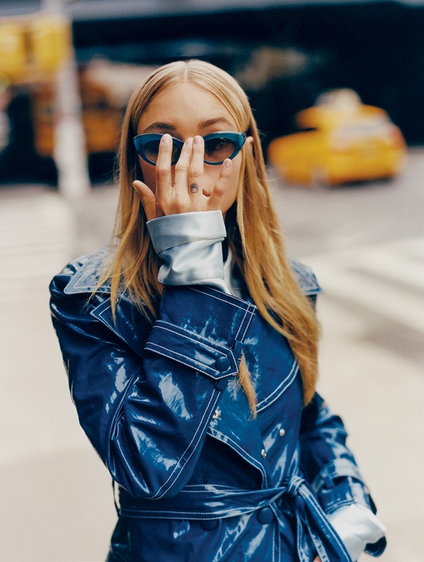 Stylish woman in blue sunglasses and a matching trench coat poses outdoors against a city backdrop with a blurred taxi.
