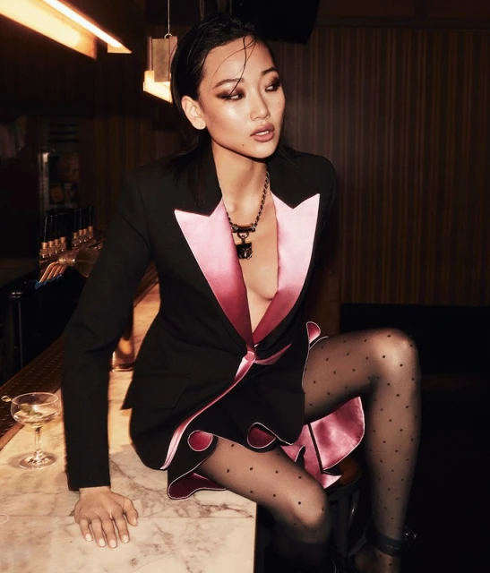 Attractive Asian woman poses in a stylish black blazer dress with pink satin trim, black patterned tights, and high heels in a bar setting.