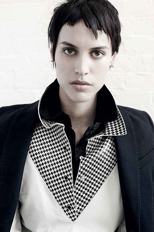 A woman poses indoors wearing a blazer, shirt, and dress pants against a white background. The subject is center framed in a portrait.