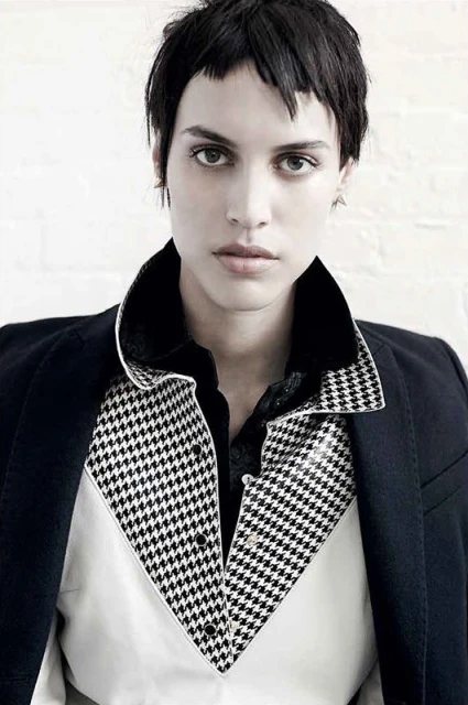 A woman poses indoors wearing a blazer, shirt, and dress pants against a white background. The subject is center framed in a portrait.