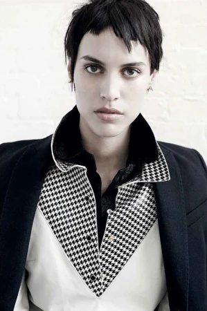A woman poses indoors wearing a blazer, shirt, and dress pants against a white background. The subject is center framed in a portrait.