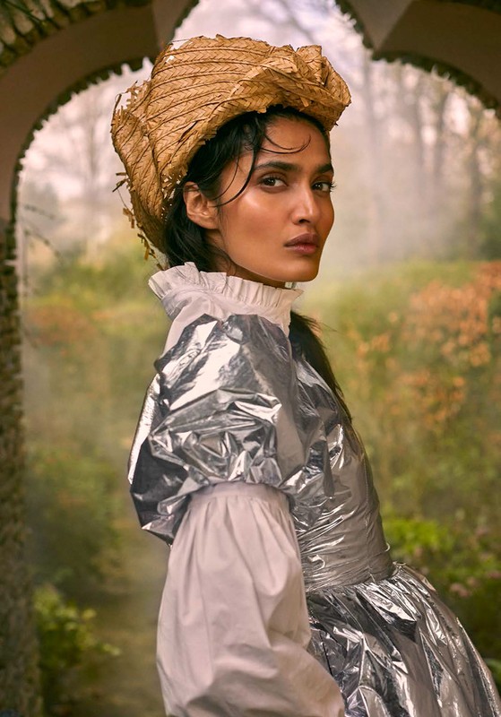 Fashionable woman in silver dress and straw hat poses in garden with archway. Elegant, high fashion, and nature-inspired visual.