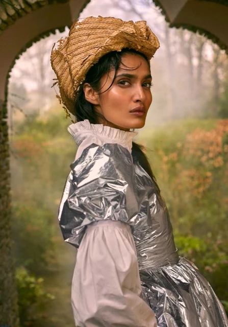 Fashionable woman in silver dress and straw hat poses in garden with archway. Elegant, high fashion, and nature-inspired visual.
