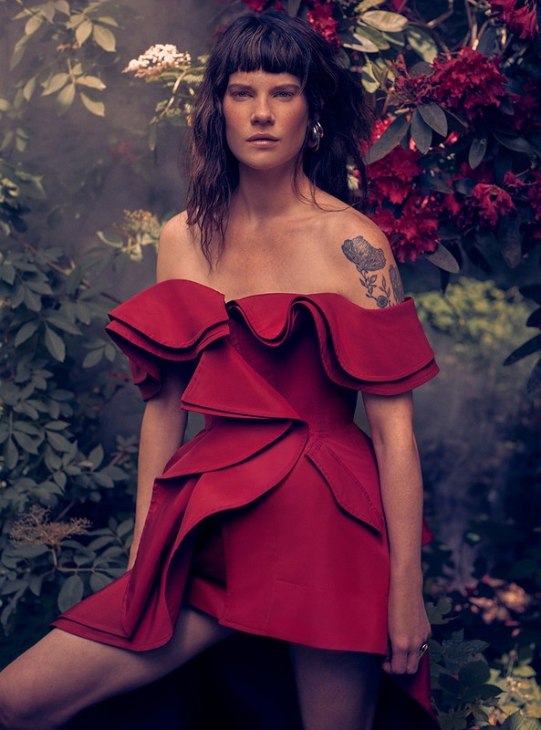 Adult female in a red, ruffled, off-the-shoulder dress poses in a lush outdoor setting with white and red flowers, and green foliage.