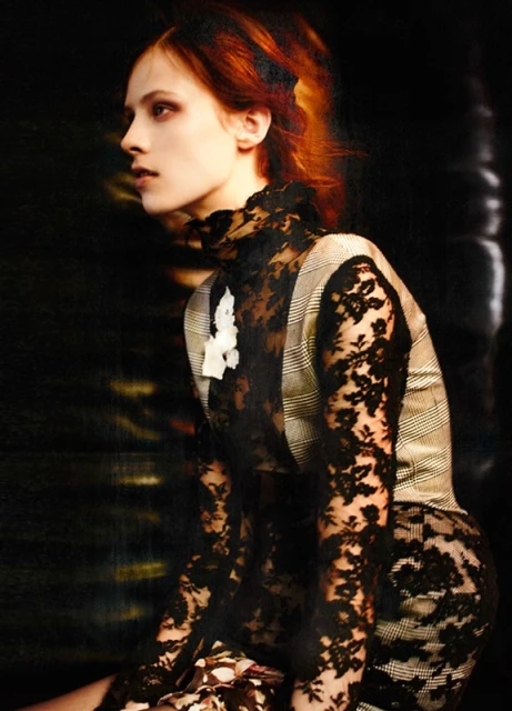 A fair-skinned woman with red hair poses in a black lace top and plaid vest against a dark background, showcasing a vintage fashion look.
