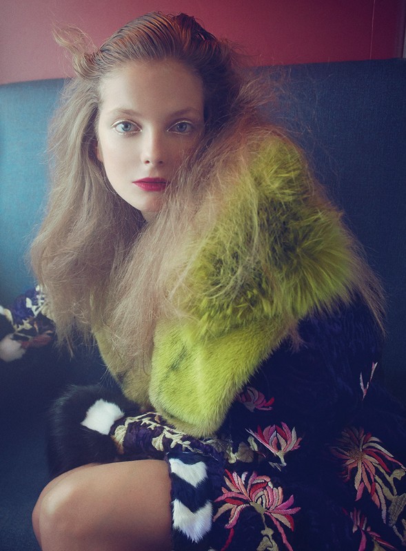 Portrait of a fair-skinned woman with fair hair and red lips wearing a dark floral dress and a light green fur jacket indoors.