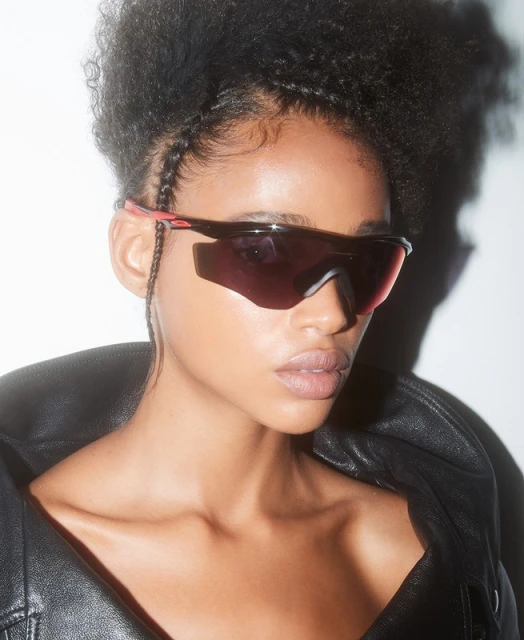 Close-up of a woman with an afro wearing sport sunglasses and a black leather jacket in front of a white backdrop.