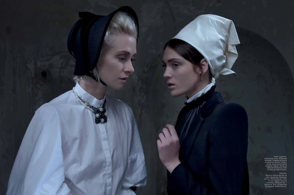 Two women wearing vintage bonnets pose in a dimly lit space. One wears white, the other black. Dark shadows and elegant clothing.