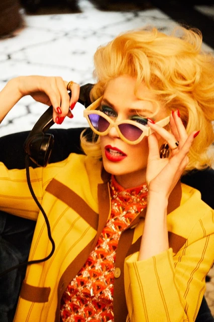 Woman with blonde hair wears sunglasses and a yellow jacket while holding an old-fashioned telephone receiver indoors.