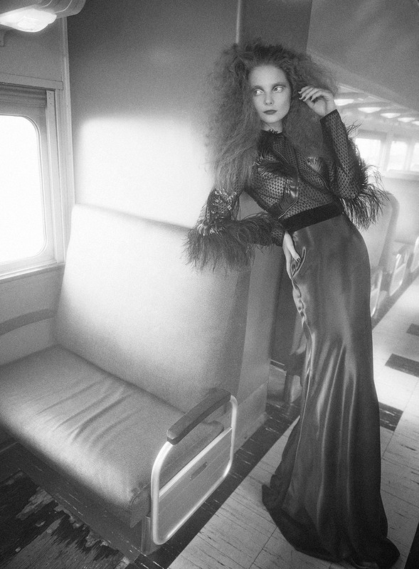 A woman with frizzy hair poses in a sheer black top and a long skirt in a vintage train car setting, with soft lighting.