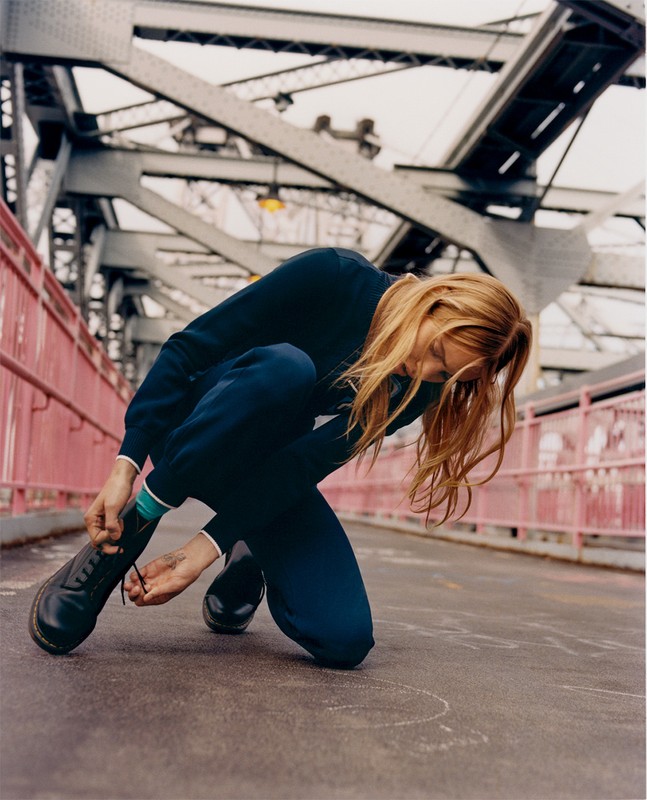 Woman in blue tracksuit laces up a black shoe while crouching on a bridge with metal supports and pink railings on an overcast day.