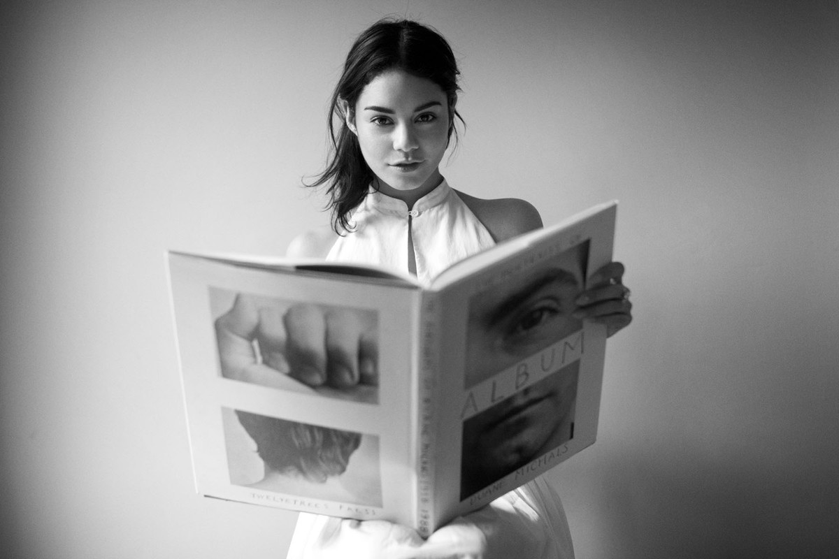 Monochrome portrait of a young woman indoors, looking at the camera and holding open a book with images of faces and torsos.