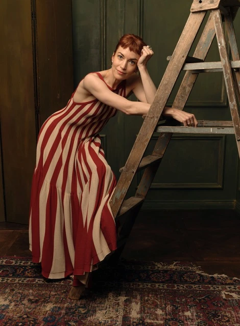 Woman in striped dress poses by wooden ladder against dark green wall, indoor shot. Fashion editorial with studio lighting.