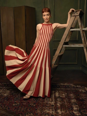 Woman in a red and cream striped halter dress leaning against a wooden ladder in a studio with green walls and a patterned rug.