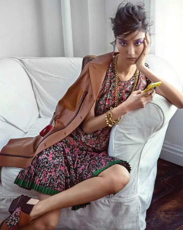 Model with purple eyeshadow sitting on a white sofa wearing a floral dress and brown jacket
