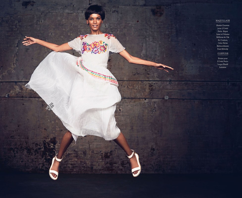 Fashion shot of an African-American woman jumping in the air wearing a white dress with floral embroidery and ankle-strap heels against a gray background.