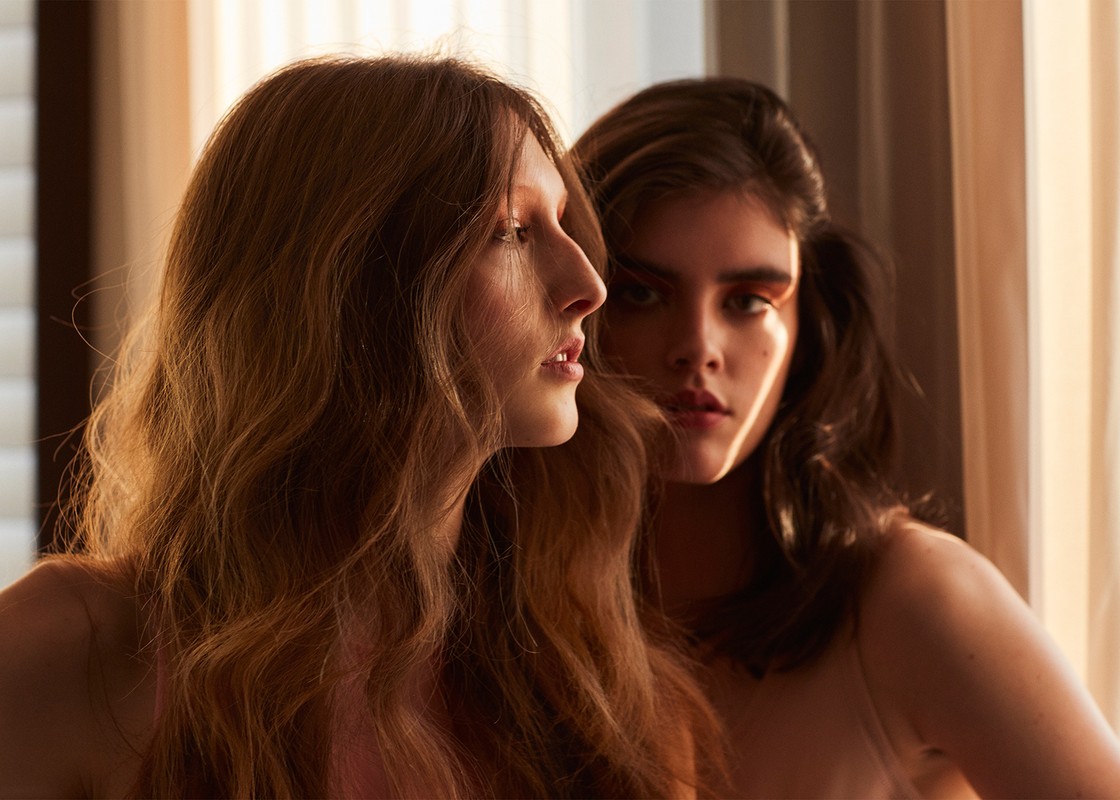 Two young women pose indoors with soft, warm lighting. The focus is on their faces and hair, creating a fashion-oriented, intimate portrait.