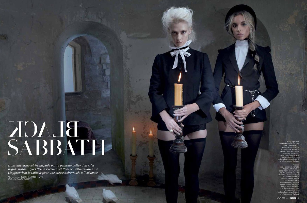 Two female models in black suits hold candles in a dark room with stone walls. Gothic fashion with an old world, elegant aesthetic.