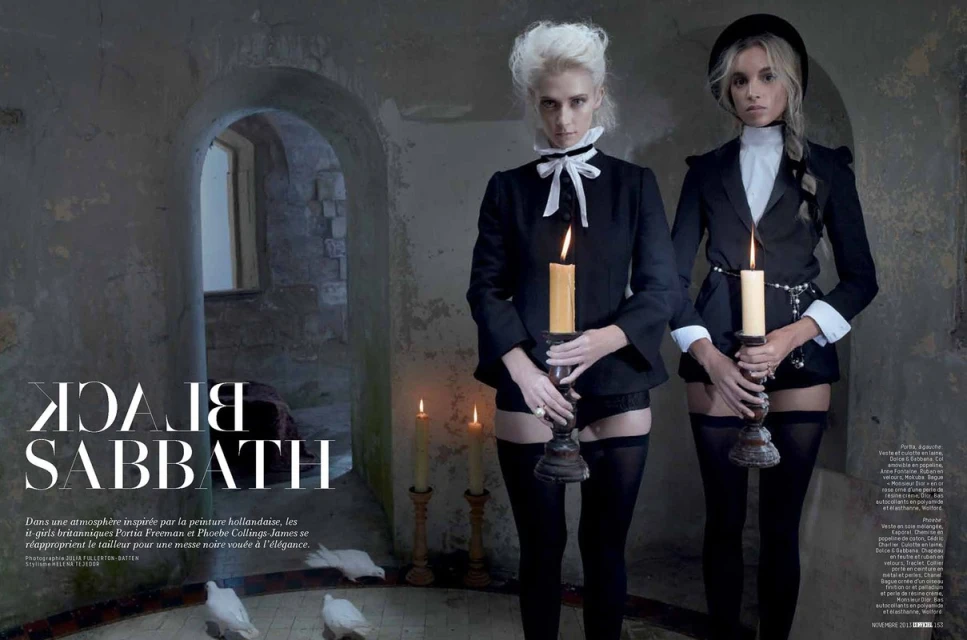 Two female models in black suits hold candles in a dark room with stone walls. Gothic fashion with an old world, elegant aesthetic.