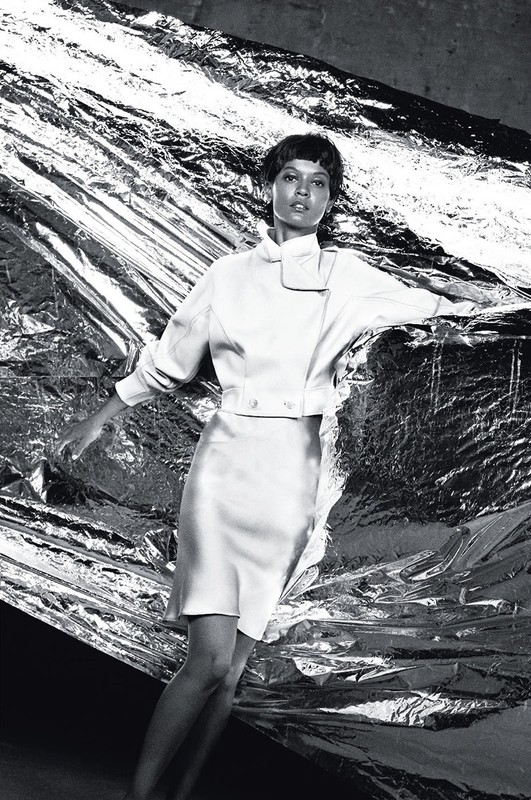 Black and white studio fashion shot of an adult female model posing with an open white jacket and skirt against a silver foil background.