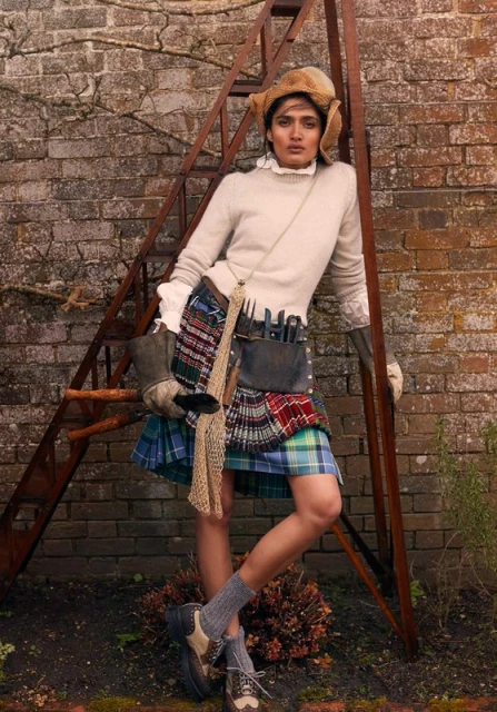 Woman in an autumn garden wearing a kilt, sweater, and fur hat stands against a rustic brick wall and leaning on an iron ladder.