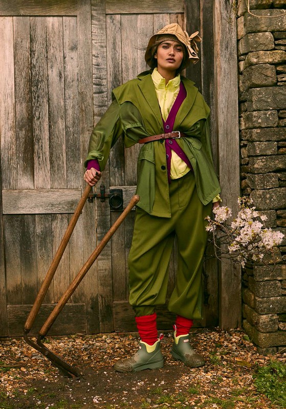 A woman in a vintage green suit and hat poses with garden tools in front of a weathered wooden door with a stone wall backdrop.