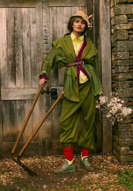 A woman in a vintage green suit and hat poses with garden tools in front of a weathered wooden door with a stone wall backdrop.