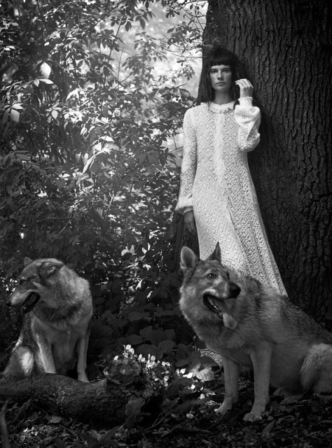 Monochrome outdoor fashion shot: woman in white lace dress with two wolfdogs in wooded setting with trees and dappled light.
