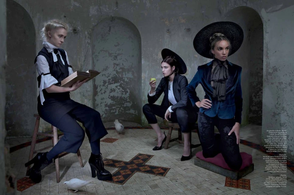 Editorial fashion shoot in an aged interior features three female models in stylish dark suits, black hats and white doves. 