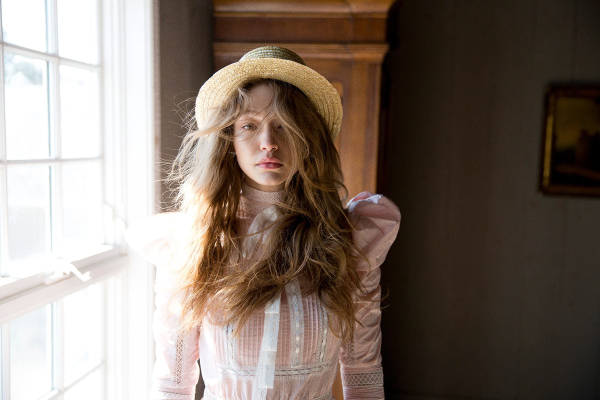 Woman in a pink Victorian-style dress and straw boater hat standing indoors near a bright window with hair styling by Kenna.