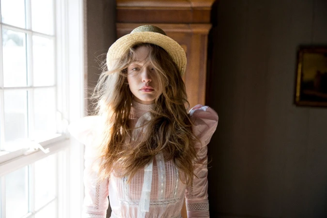 Woman in a pink Victorian-style dress and straw boater hat standing indoors near a bright window with hair styling by Kenna.