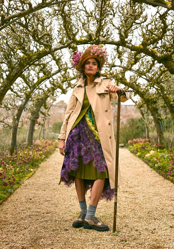 Woman in trench coat and floral hat poses on garden path under flower archway, holding a walking stick, in an outdoor fashion portrait.
