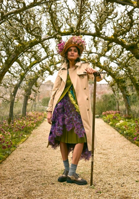 Woman in trench coat and floral hat poses on garden path under flower archway, holding a walking stick, in an outdoor fashion portrait.