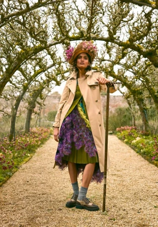 Woman in trench coat and floral hat poses on garden path under flower archway, holding a walking stick, in an outdoor fashion portrait.