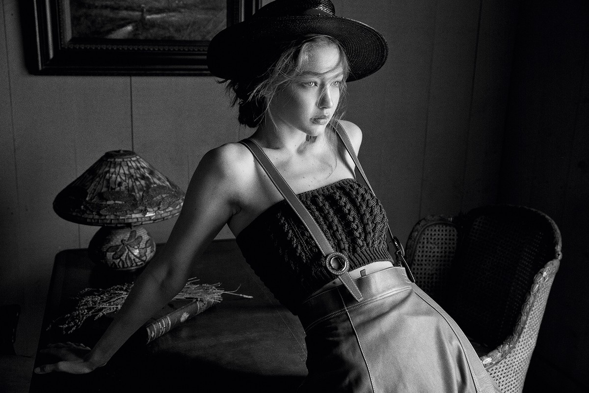 Monochrome portrait of a woman wearing a hat and a knit top, leaning on a table next to a lamp, in a dimly lit room.