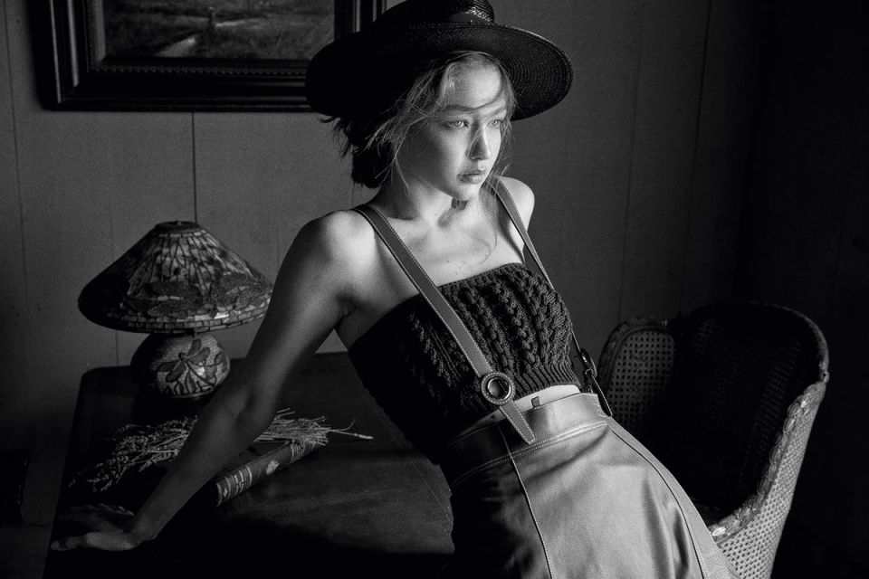 Monochrome portrait of a woman wearing a hat and a knit top, leaning on a table next to a lamp, in a dimly lit room.