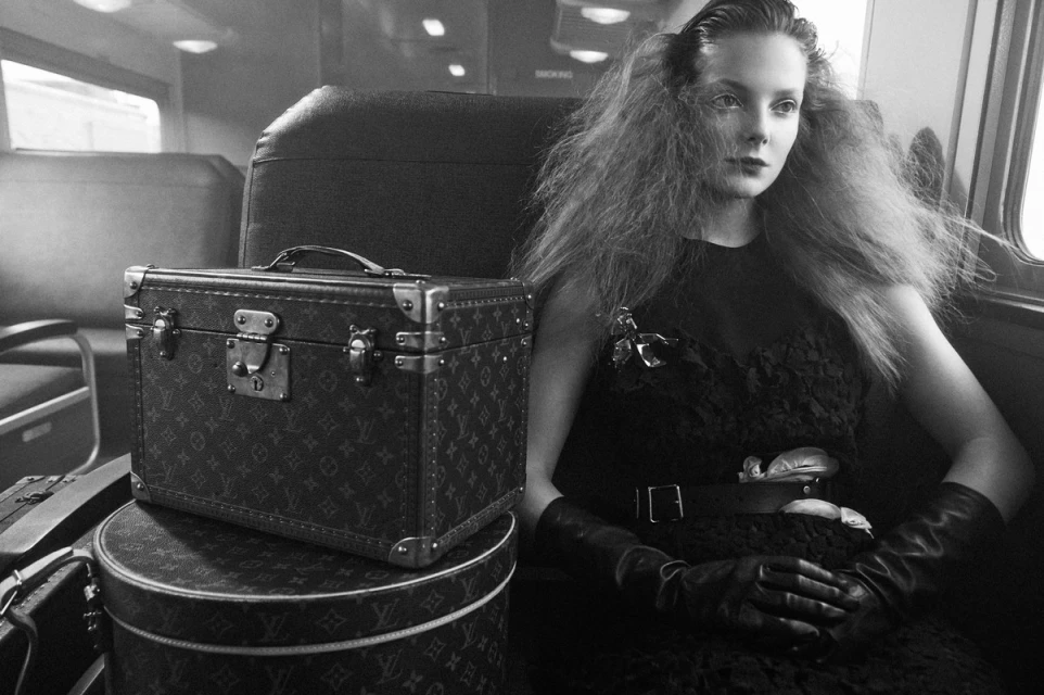 Black and white image of a woman with textured hair sitting on a train next to vintage Louis Vuitton luggage with a classic, neutral tone.