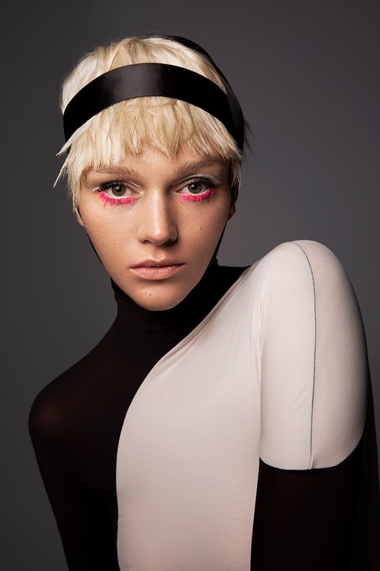 Close-up portrait of a woman with short blonde hair, black headband, and pink eye makeup against a plain gray background in a studio.