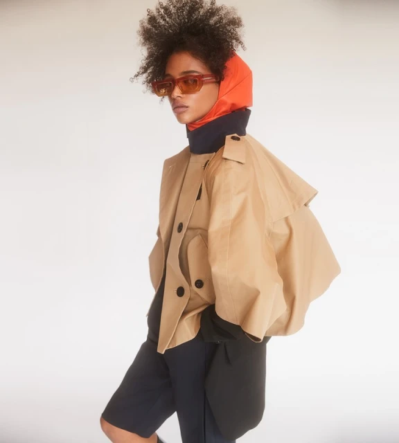 Woman in orange hood and sunglasses wearing a tan trench coat in a studio portrait with a light background for a fashion editorial.